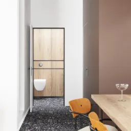 Nowoczesna łazienka z ukrytą toaletą za drewnianą ścianą, widok z jadalni z drewnianym stołem i krzesłami na tle podłogi z lastryko.