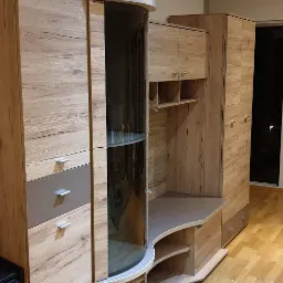 Zmontowana, nowoczesna meblościanka z forniru imitującego drewno, z przeszkloną witryną i półkami, na jasnej podłodze z paneli.