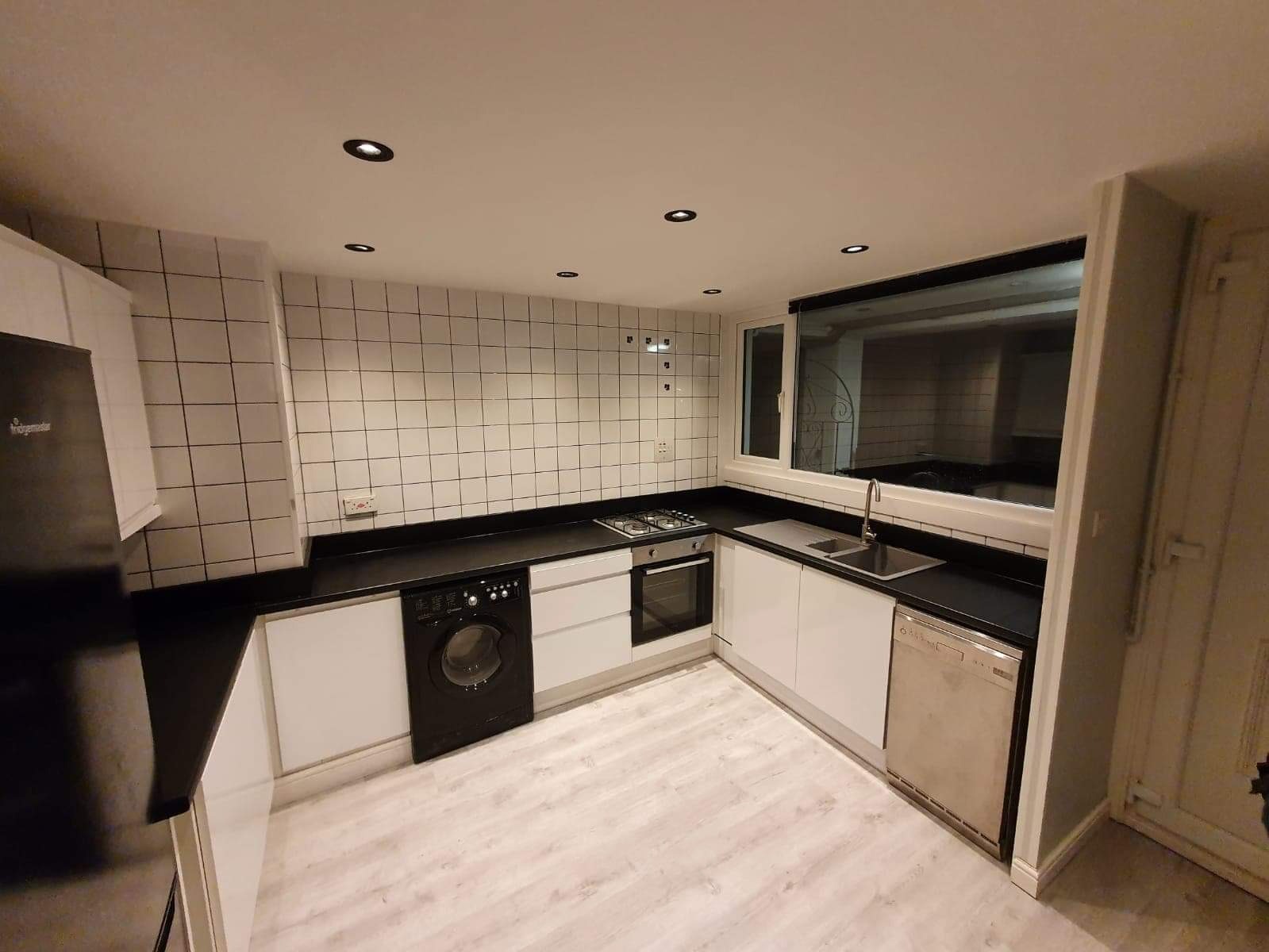 Nowoczesna kuchnia z białymi szafkami i czarnym blatem, wykończona białymi kwadratowymi płytkami, z widoczną pralką w zabudowie i drewnopodobną podłogą.