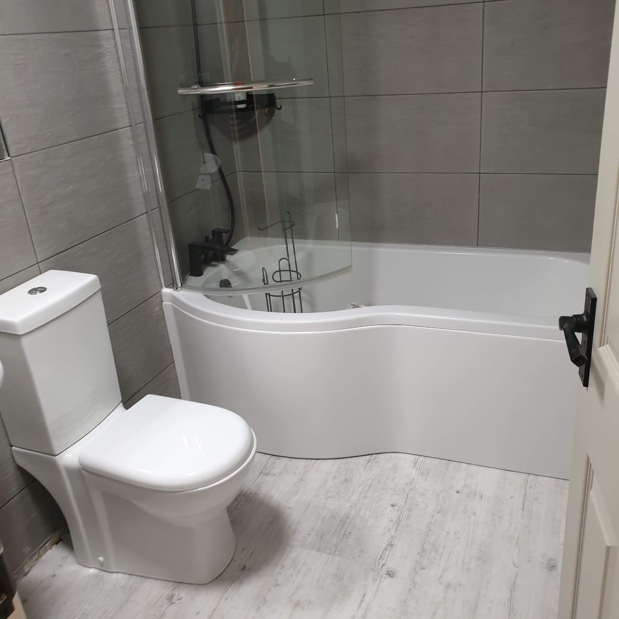 Nowoczesna łazienka w szaro-białej tonacji z płytkami imitującymi kamień, białą wanną z przeszklonym parawanem nawannowym, wiszącą toaletą i podłogą z paneli winylowych imitujących jasne drewno.