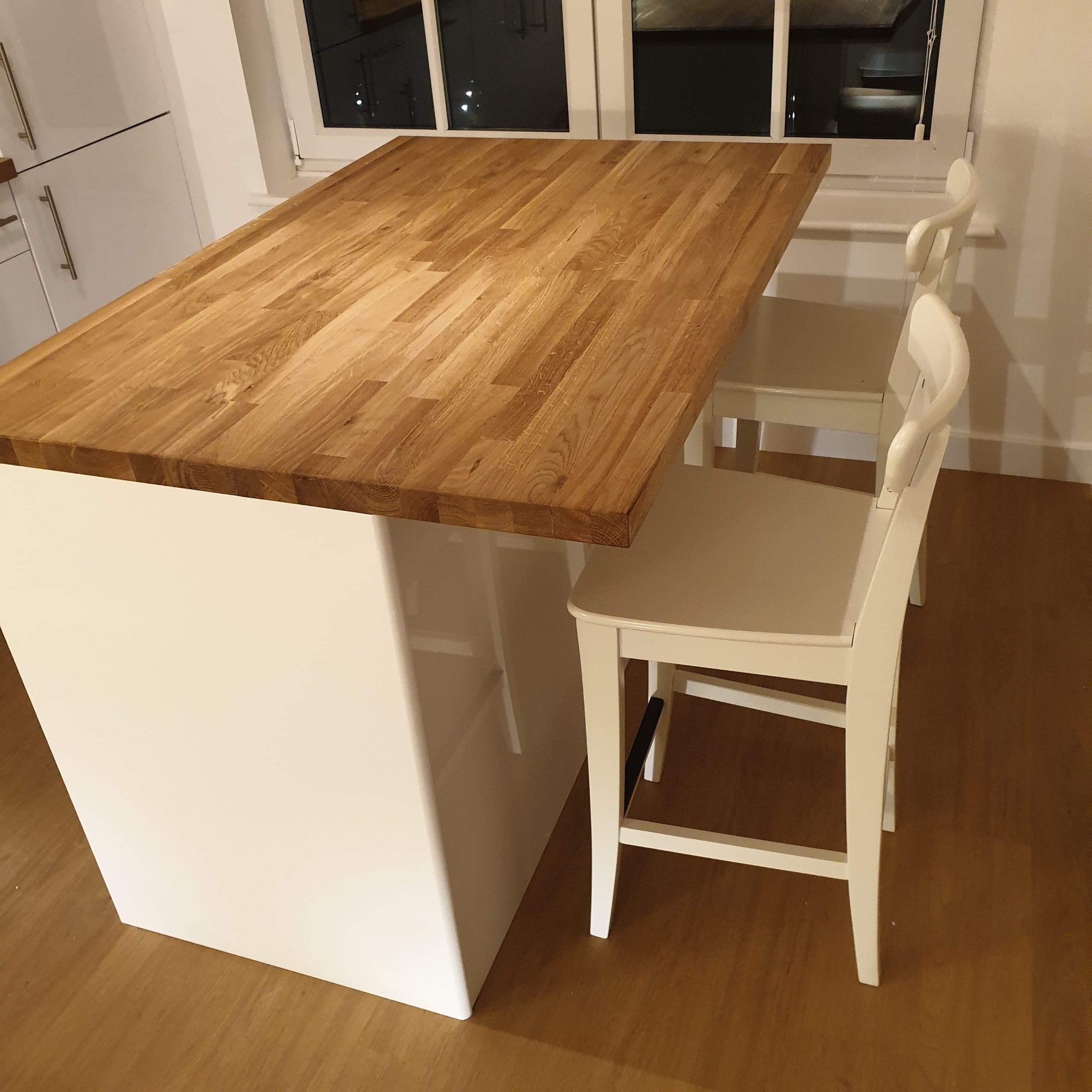 Wysoka wyspa kuchenna z drewnianym blatem i białymi szafkami, uzupełniona dwoma białymi krzesłami barowymi na tle okna.