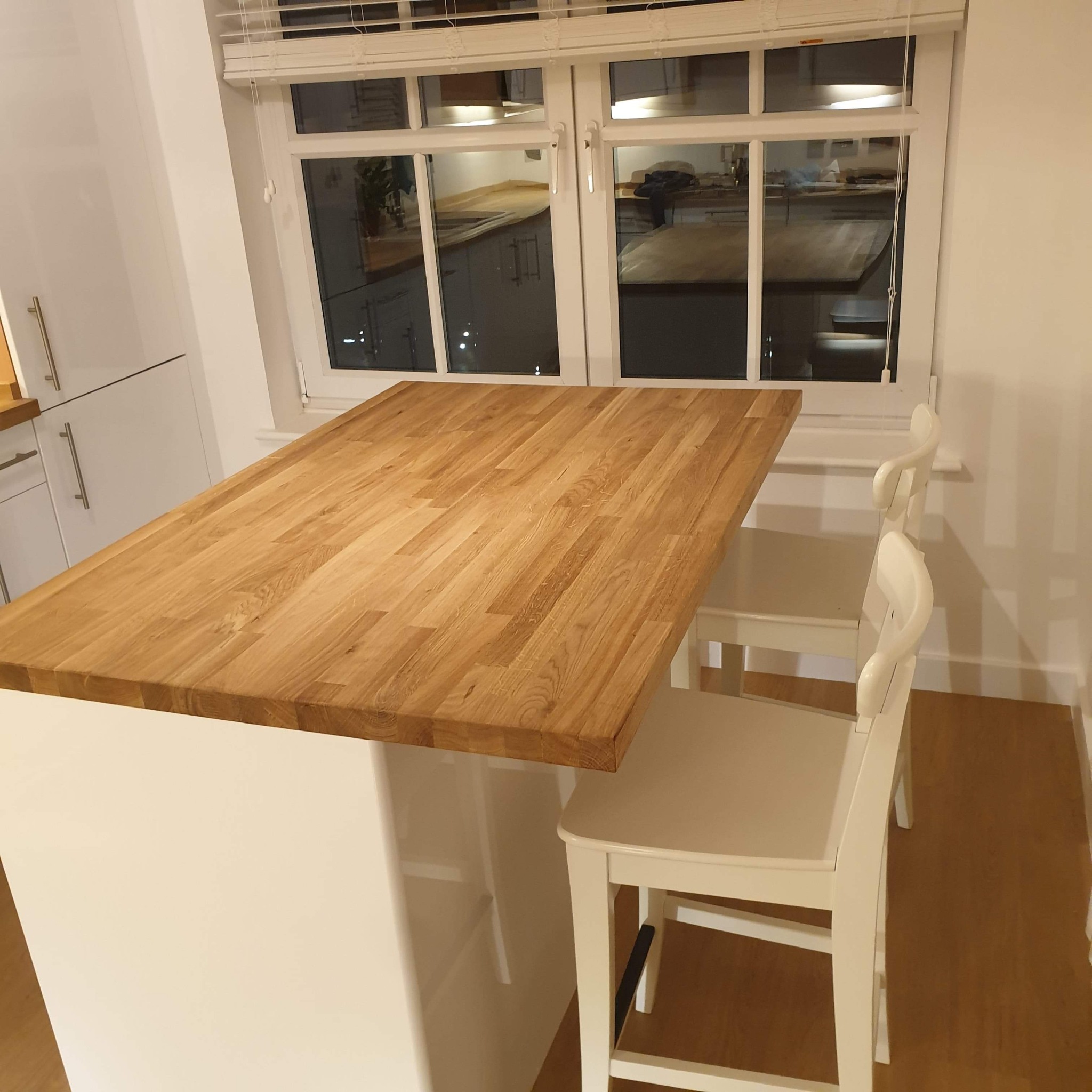 Drewniany blat wyspy kuchennej zamontowany na białej szafce, widoczne dwa białe krzesła barowe i okno z żaluzjami.