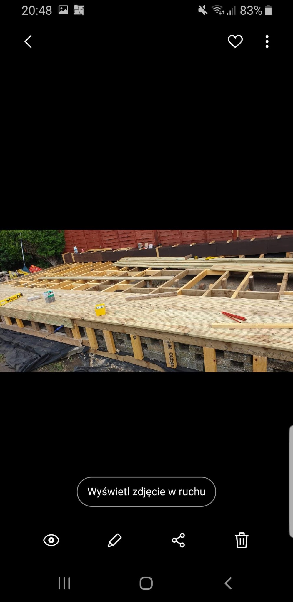 Konstrukcja drewnianego tarasu na podwyższeniu, widoczne legary i deski, poziomica, skrzynka z wkrętami, ogrodzenie w tle.