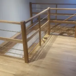 Drewniana balustrada z metalowymi prętami na antresoli, widoczne narzędzia montażowe, podłoga laminowana w odcieniach beżu.