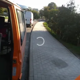 Świeżo ułożona szara kostka brukowa tworząca chodnik, widziana z perspektywy zaparkowanego pomarańczowego busa, z białym dostawczakiem w tle i zielenią drzew w oddali.