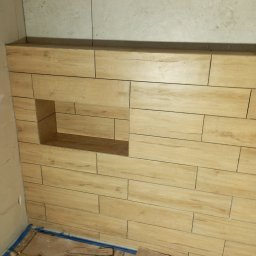 Profesjonalne usługi malarskie i remonty wnętrz. - Ściana wyłożona podłużnymi, drewnopodobnymi płytkami z wnęką. U góry ściana wyłożona jasnymi, kwadratowymi płytkami. Na podłodze zabezpieczenie z kartonu.