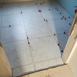 Profesjonalne usługi malarskie i remonty wnętrz. - Jasne płytki podłogowe w trakcie układania, z pomarańczowymi klinami poziomującymi. Widok z góry na fragment łazienki z surowymi ścianami.