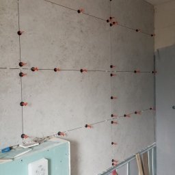 Profesjonalne usługi malarskie i remonty wnętrz. - Ściana w trakcie układania płytek, widoczne systemy poziomowania płytek (czarne klipsy i pomarańczowe kliny), sznurki wyznaczające linie. Fragment ściany z płyty G-K.