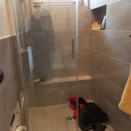 Profesjonalne usługi malarskie i remonty wnętrz. - Wykończona kabina prysznicowa z szarymi płytkami imitującymi marmur, z widoczną pralką z lewej strony. W kabinie leżą ubrania.