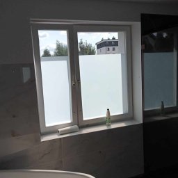 Profesjonalne usługi malarskie i remonty wnętrz. - Okno w łazience z naklejoną folią matującą, częściowo widoczna wanna. Na parapecie rolka folii i butelka. Na zewnątrz widok na budynek i drzewa.