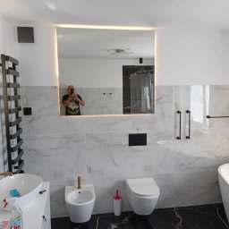 Profesjonalne usługi malarskie i remonty wnętrz. - Elegancka łazienka w bieli i czerni z marmurowymi płytkami i podświetlanym lustrem. Widoczna armatura w kolorze miedzi i czarne dodatki. W lustrze odbicie osoby robiącej zdjęcie.