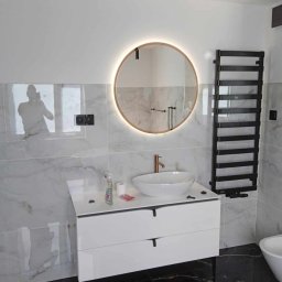 Profesjonalne usługi malarskie i remonty wnętrz. - Nowoczesna łazienka po metamorfozie: białe płytki imitujące marmur, okrągłe lustro z podświetleniem LED i czarne akcenty. Widoczny grzejnik drabinkowy.