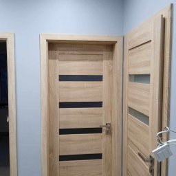 Profesjonalne usługi malarskie i remonty wnętrz. - Dwie drewniane, nowoczesne drzwi wewnętrzne z przeszkleniami, jedna zamknięta, druga otwarta, na tle szarej ściany. Widoczne detale klamek i ościeżnic.