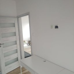 Profesjonalne usługi malarskie i remonty wnętrz. - Nowoczesny pokój po remoncie: białe ściany, drewniana podłoga, drzwi z przeszkleniami i minimalistyczne biurko. Widoczne gniazdka elektryczne.