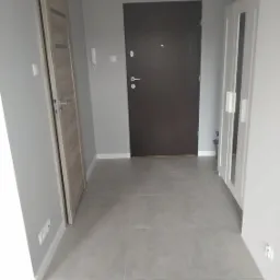 Szare płytki podłogowe w przedpokoju po remoncie, widoczne drzwi wejściowe i wewnętrzne, biała szafa z lustrem. Świeżo pomalowane ściany na szaro.