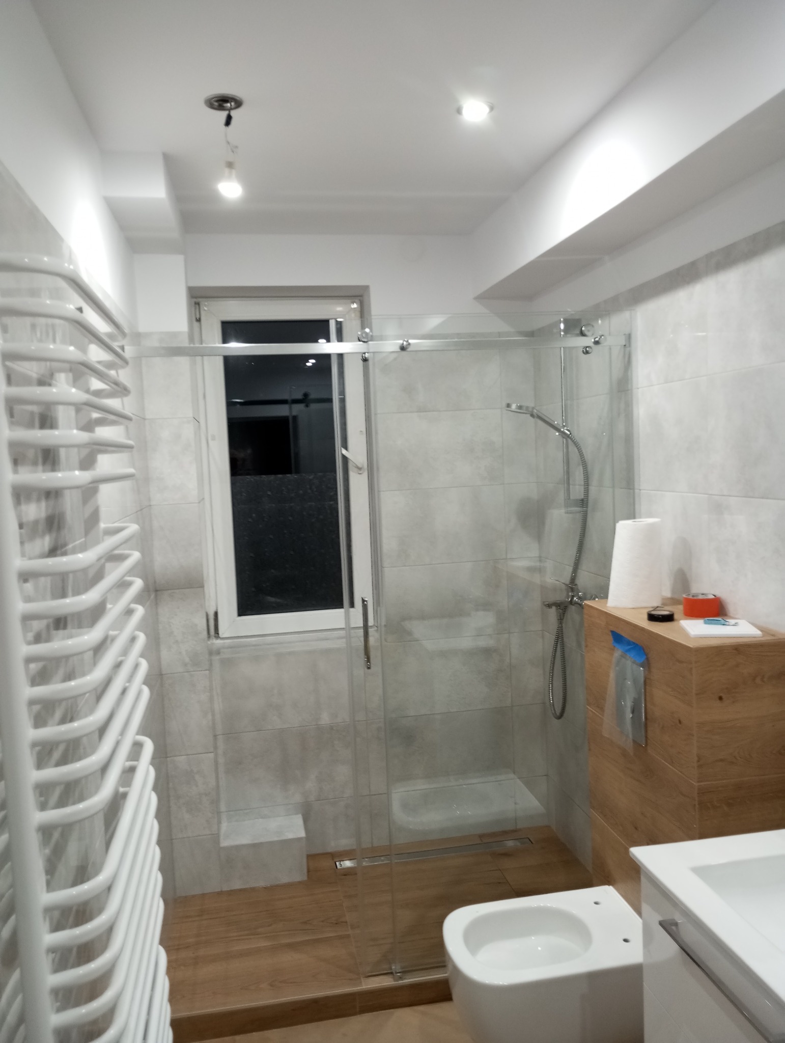 Wykończona łazienka z kabiną prysznicową ze szklanymi drzwiami przesuwnymi, grzejnikiem drabinkowym i drewnopodobną podłogą. Widoczne szare płytki na ścianach.