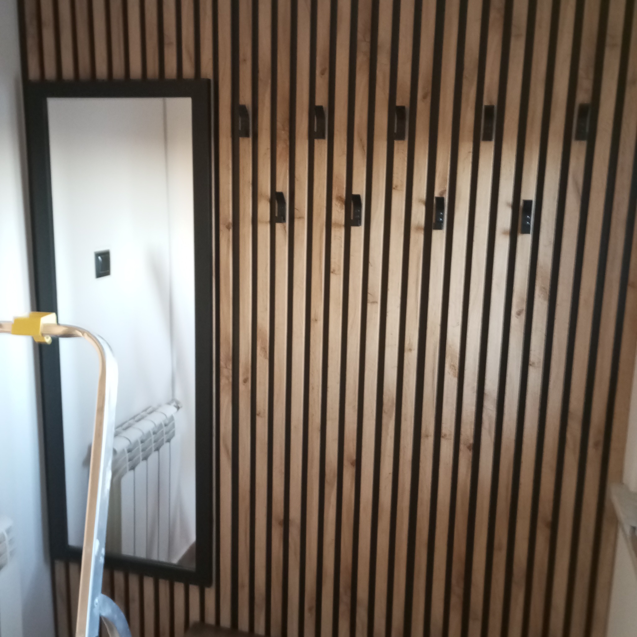 Ściana z drewnianych lameli z czarnymi haczykami i lustro z czarną ramą. Fragment drabiny malarskiej w lewym dolnym rogu.