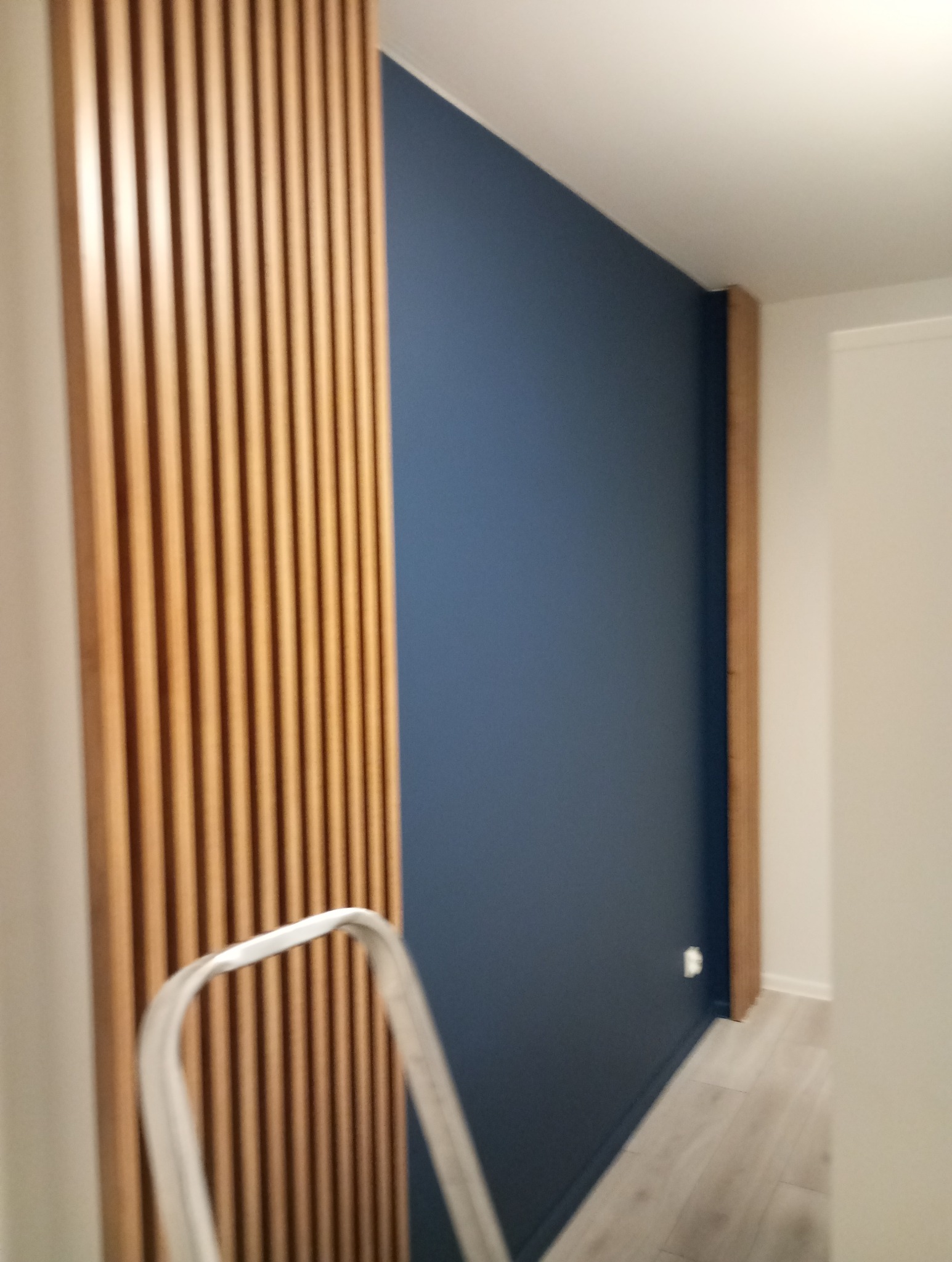 Ściana w kolorze granatowym z drewnianymi lamelami i listwą. Fragment drabiny malarskiej. Nowoczesne wykończenie wnętrza.