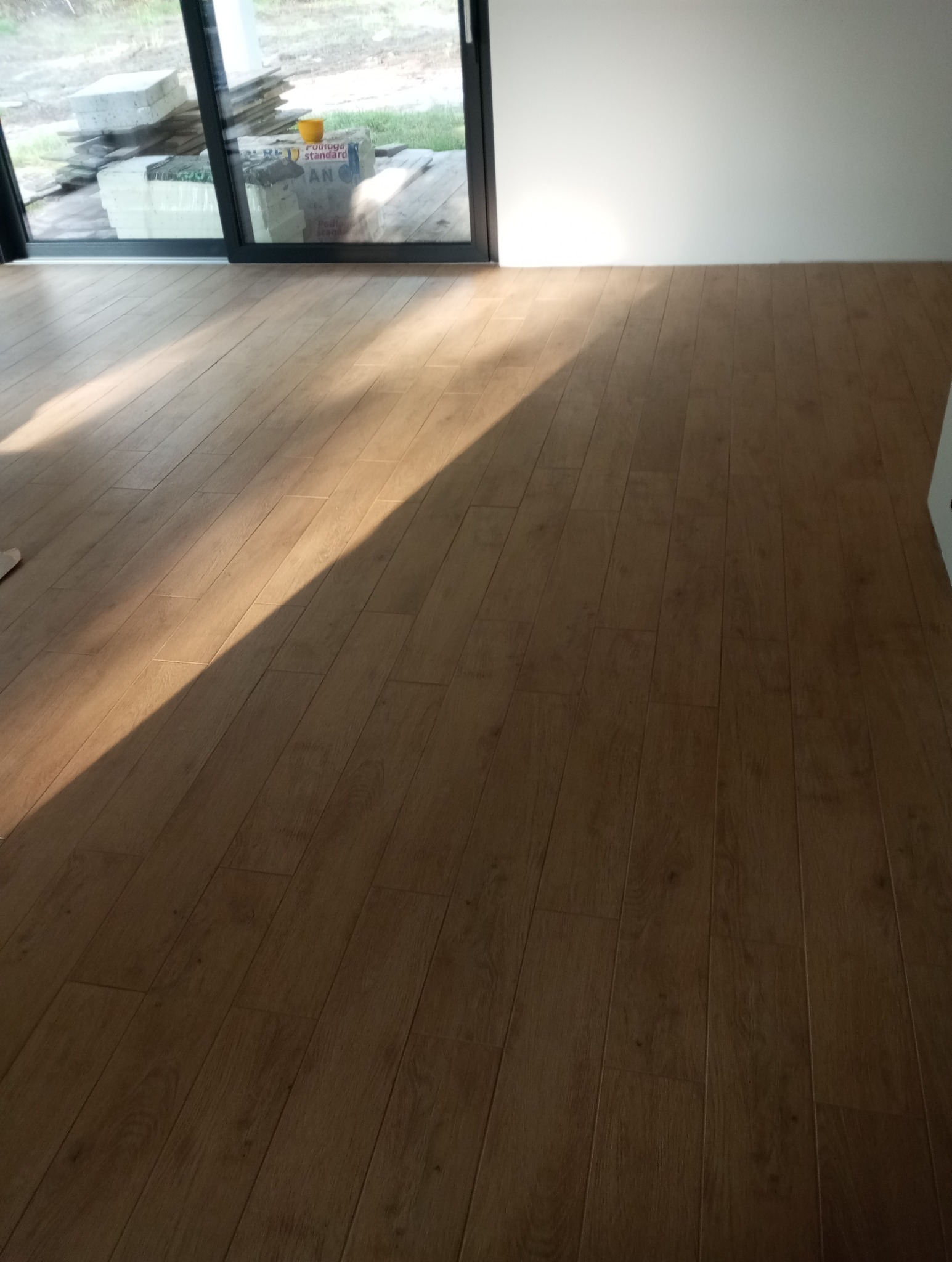 Ułożone panele podłogowe w jasnym odcieniu drewna, z widocznym padającym światłem słonecznym. W tle okno przesuwne i materiały budowlane.