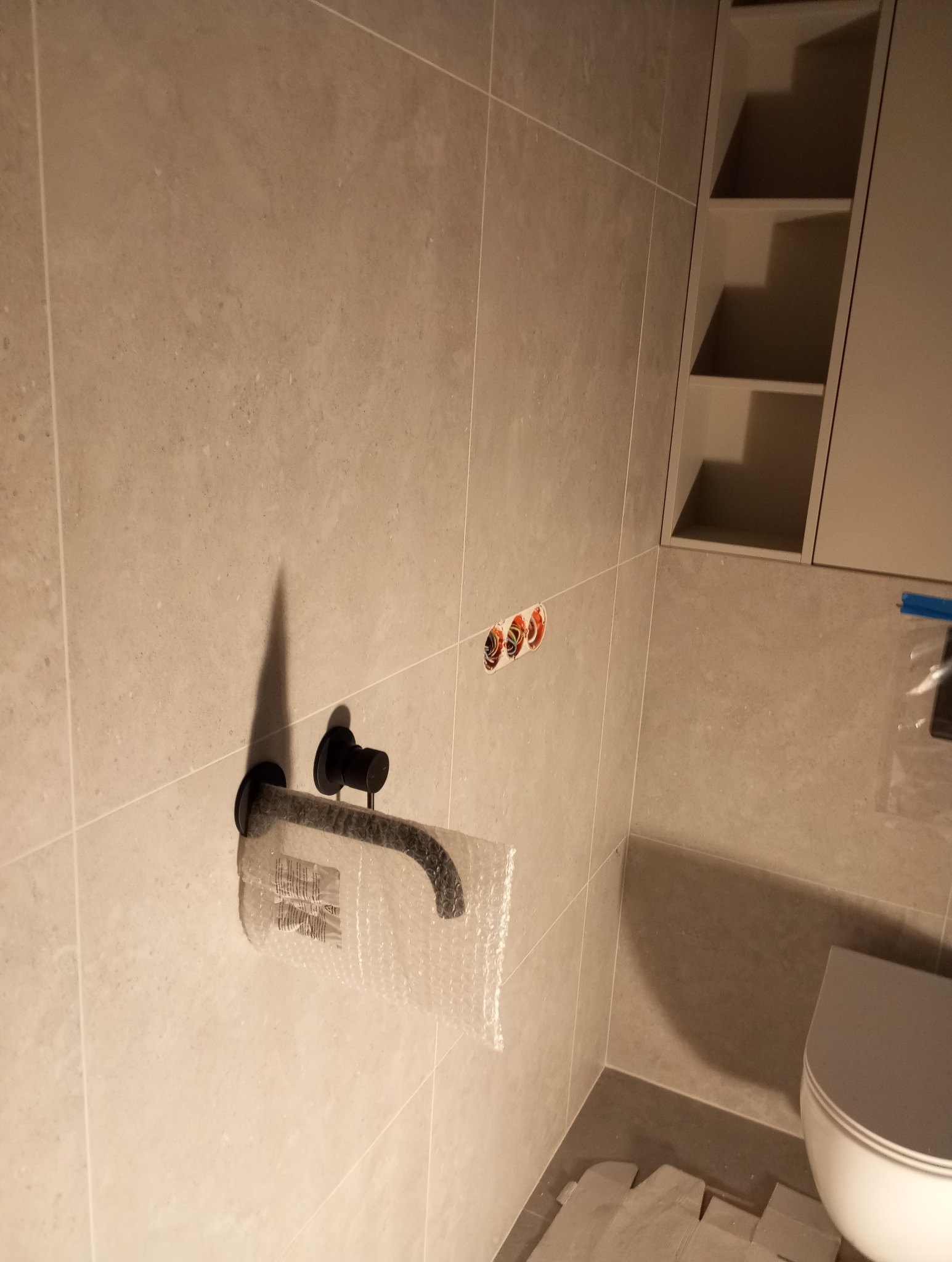 Ściana w łazience wyłożona jasnymi płytkami, z zamontowaną czarną baterią umywalkową zabezpieczoną folią bąbelkową. Widoczne przyłącza wodne i fragment zabudowy.