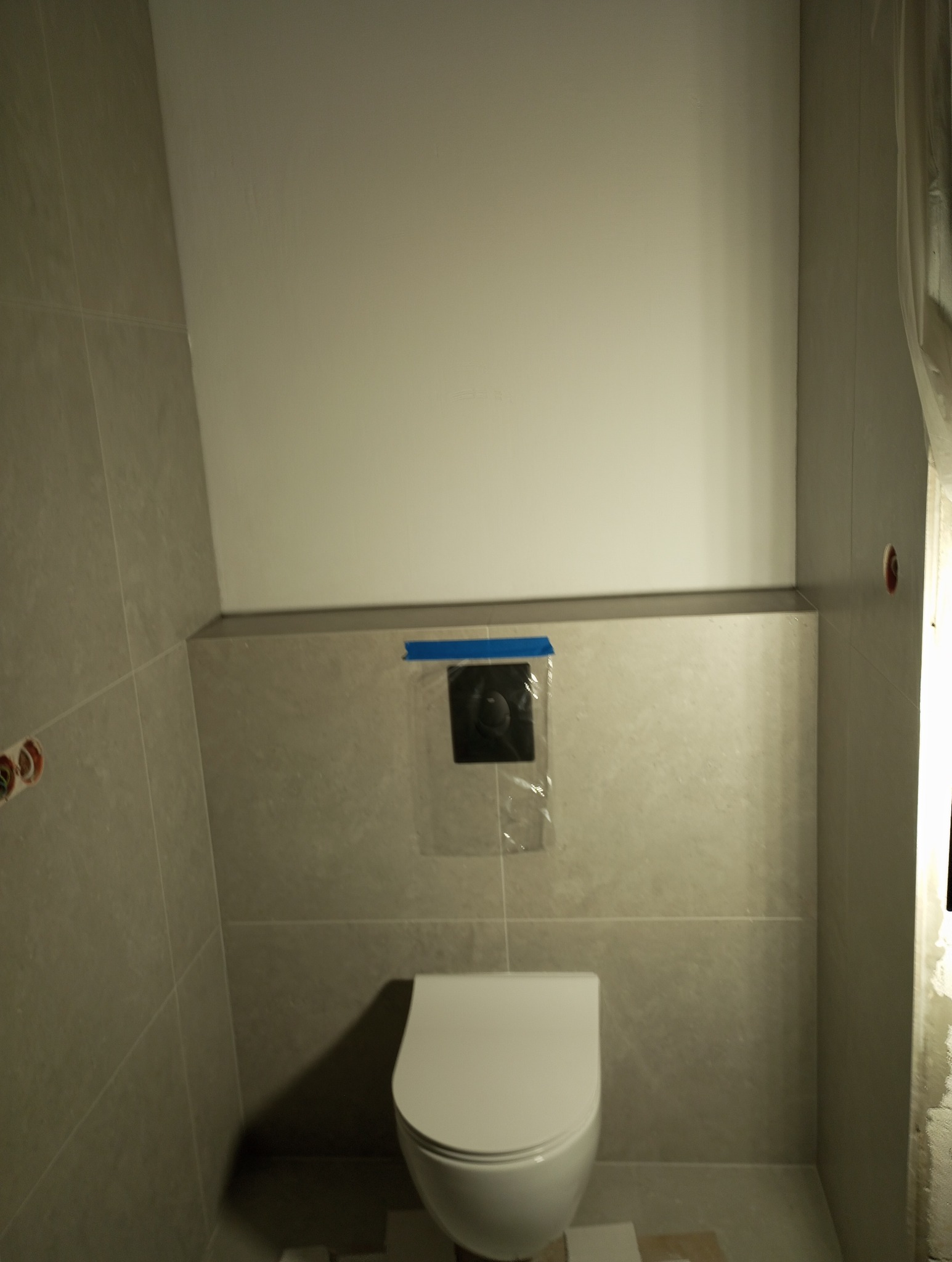 Wykończona łazienka z zamontowaną toaletą podwieszaną i szarą glazurą. Na ścianie zabezpieczony element sterujący spłuczką. Świeżo położone płytki.