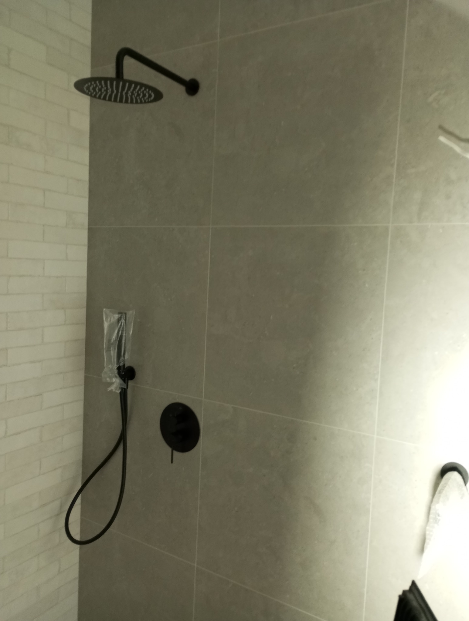 Wykończona ściana kabiny prysznicowej z szarych płytek, z czarną deszczownicą i baterią. Słuchawka prysznicowa jeszcze w folii ochronnej. Fragment ściany z białej cegły.