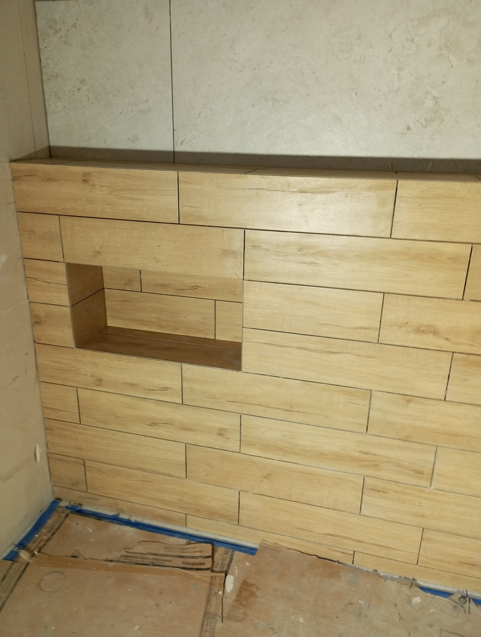 Ściana wyłożona podłużnymi, drewnopodobnymi płytkami z wnęką. U góry ściana wyłożona jasnymi, kwadratowymi płytkami. Na podłodze zabezpieczenie z kartonu.