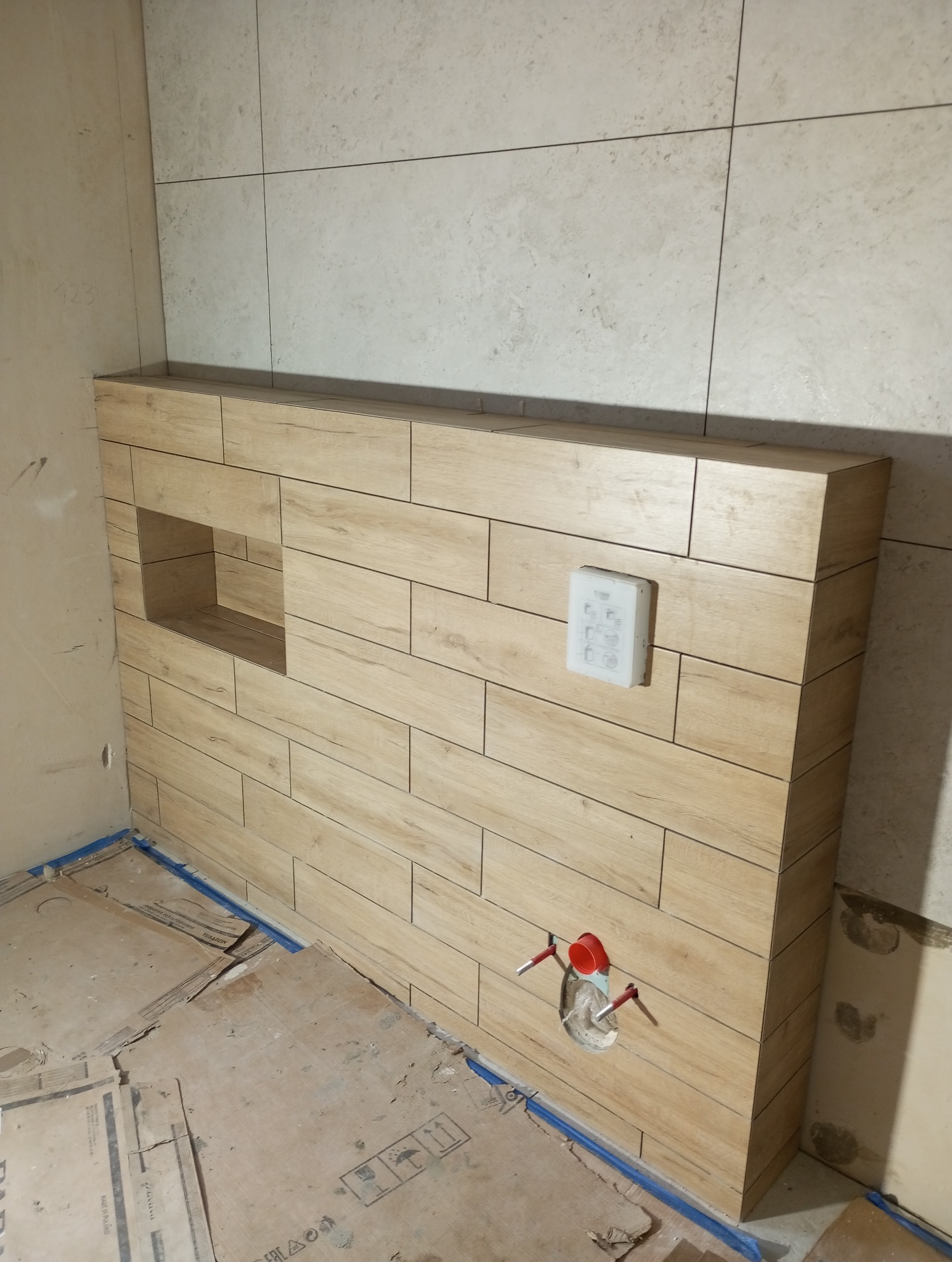 Obudowa WC z płytek imitujących drewno, z wnęką i przyciskiem spłukiwania. Widoczne rury instalacyjne. Podłoga zabezpieczona kartonem.