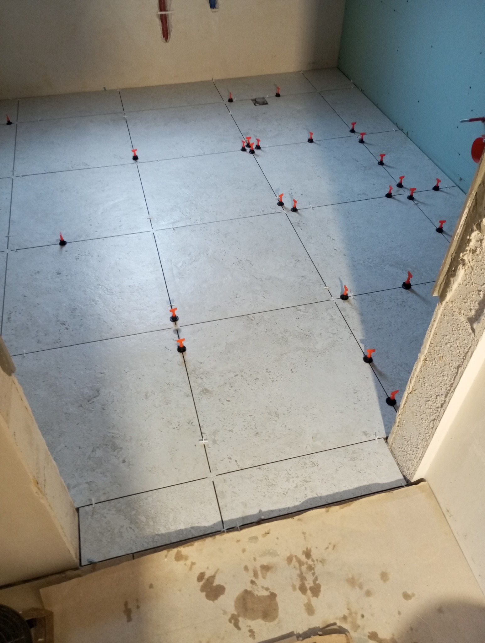 Jasne płytki podłogowe w trakcie układania, z pomarańczowymi klinami poziomującymi. Widok z góry na fragment łazienki z surowymi ścianami.