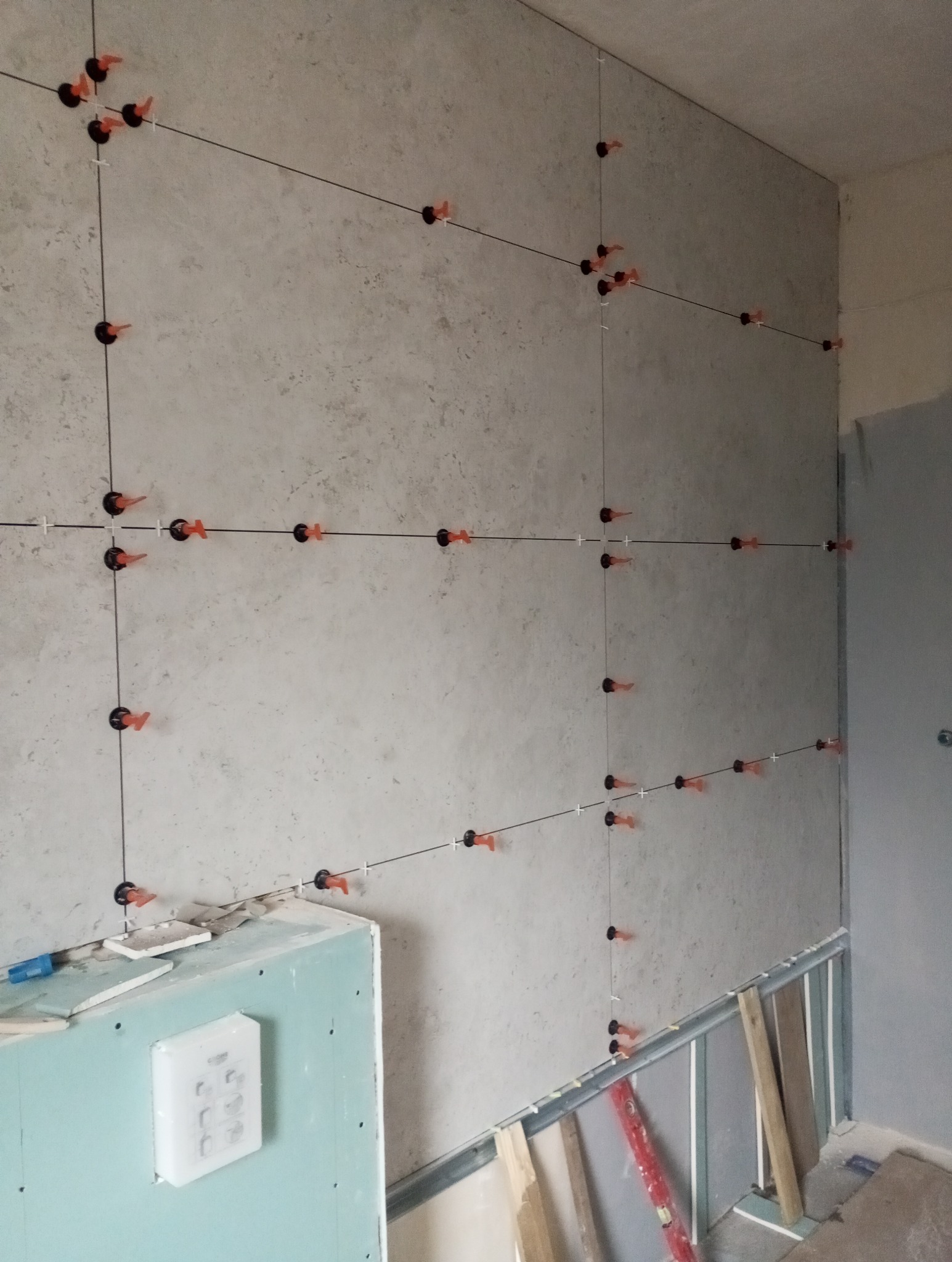 Ściana w trakcie układania płytek, widoczne systemy poziomowania płytek (czarne klipsy i pomarańczowe kliny), sznurki wyznaczające linie. Fragment ściany z płyty G-K.