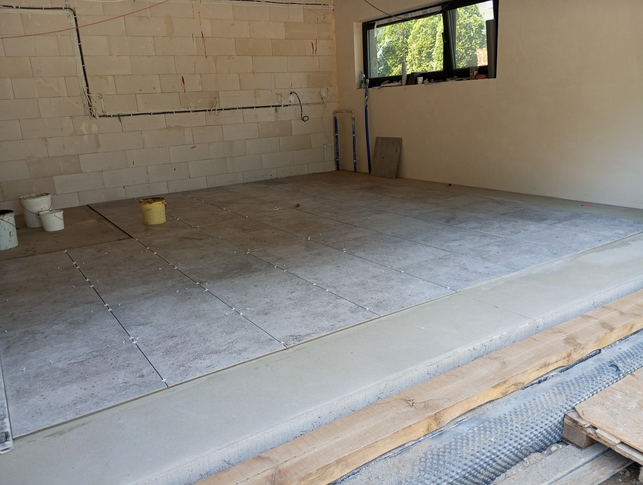 Układanie szarych płytek podłogowych w surowym wnętrzu z widocznymi przewodami i oknem. Płytki oddzielone krzyżykami dystansowymi, widoczne przygotowania podłoża.