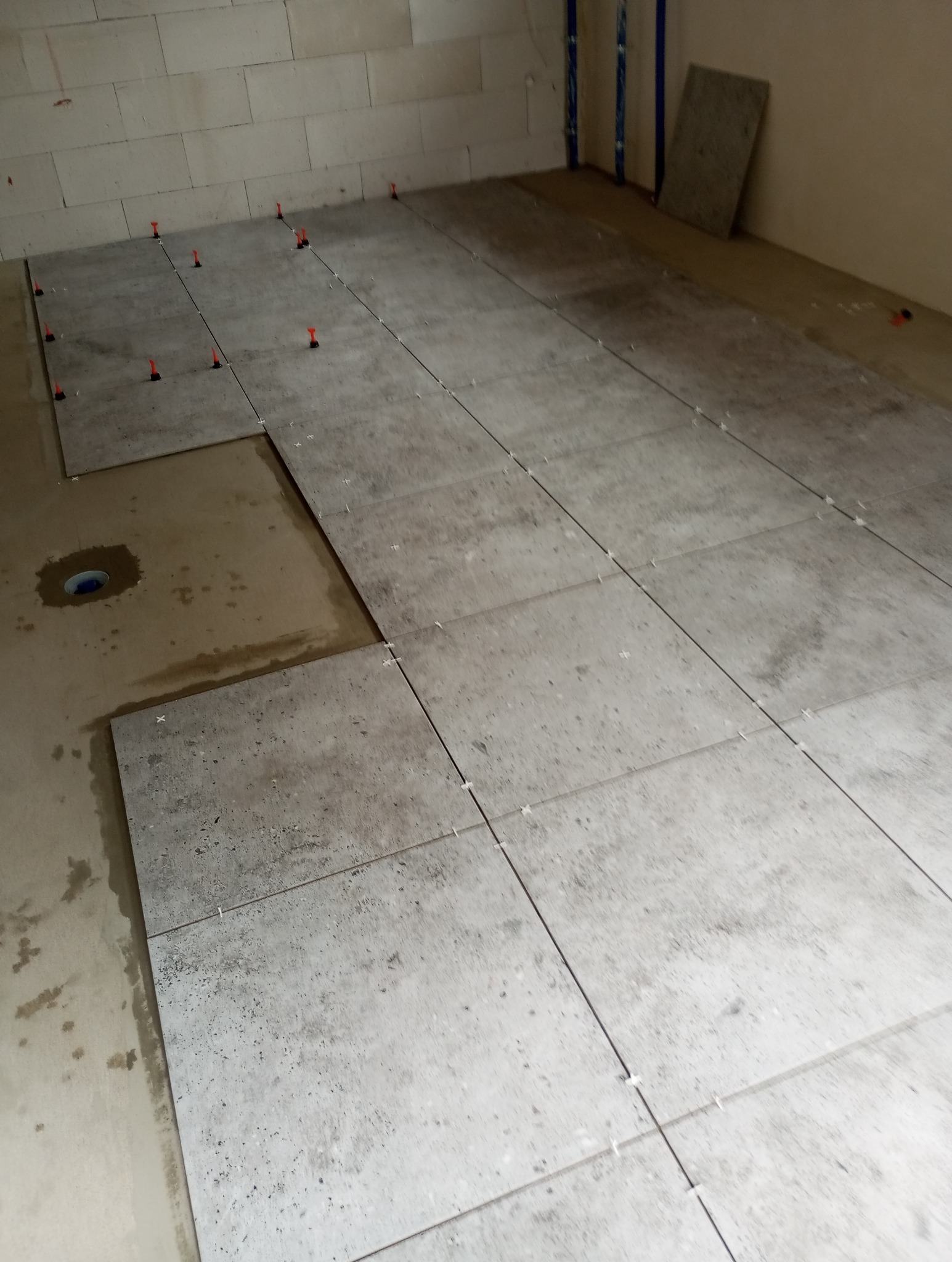 Układanie szarych, kwadratowych płytek podłogowych z użyciem poziomujących klipsów i klinów w trakcie remontu. Fragment podłogi bez płytek, widoczny odpływ.