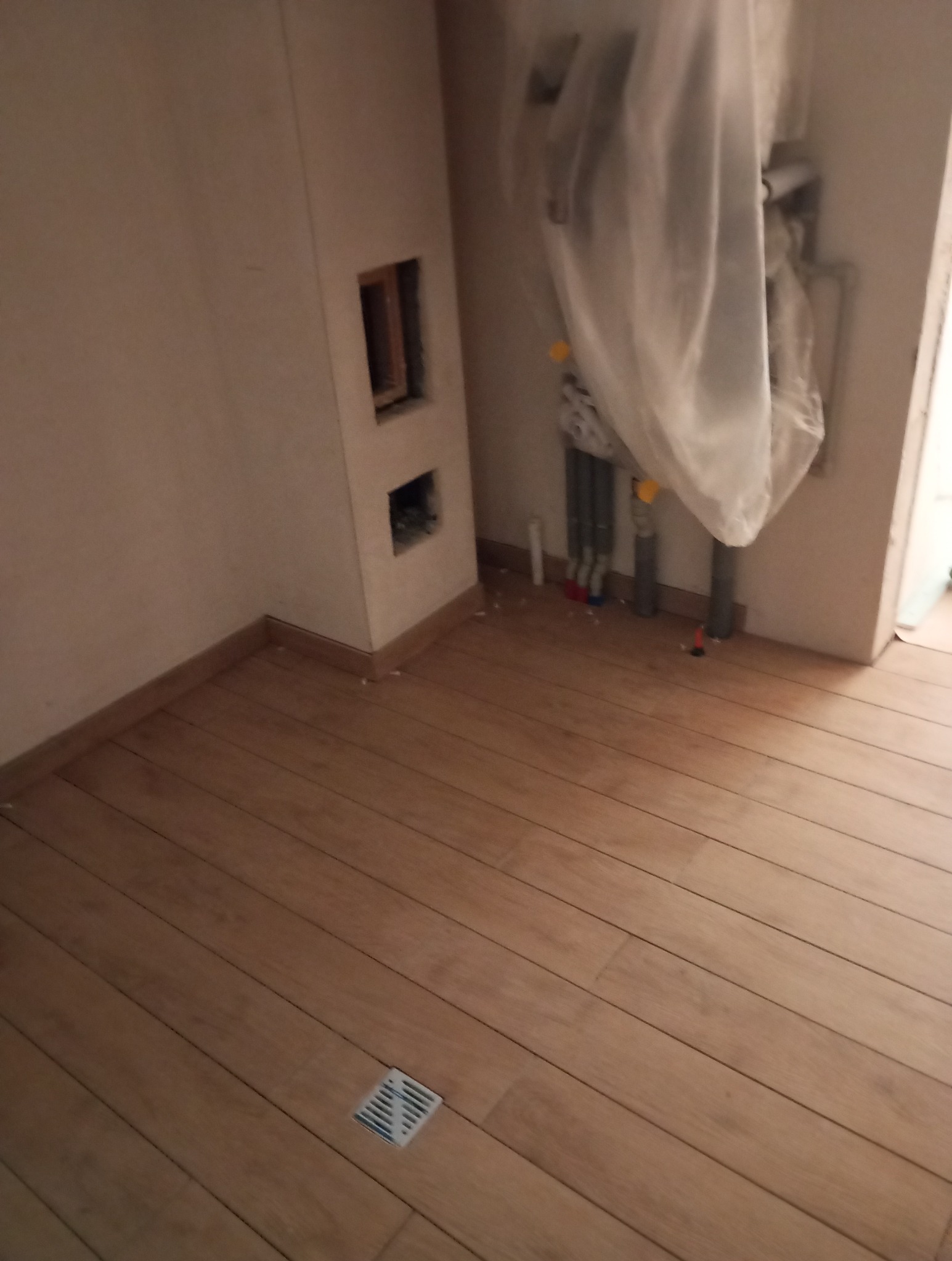 Ułożone panele podłogowe w odcieniach jasnego drewna, widoczna kratka ściekowa w podłodze, ściana z otworami instalacyjnymi i rury osłonięte folią.