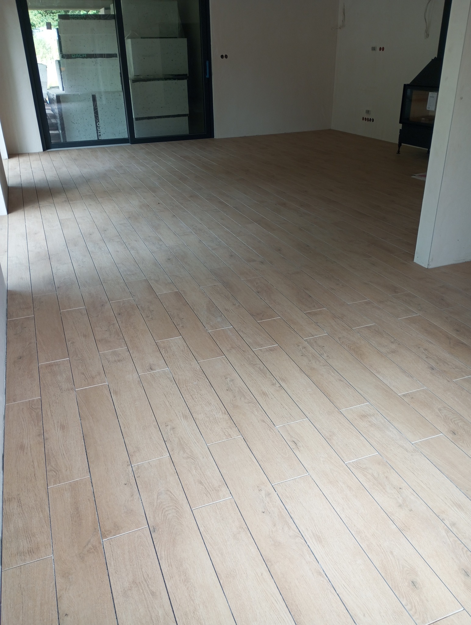 Ułożona jasna podłoga z paneli imitujących drewno w nowym wnętrzu. Widoczny fragment kominka i duże okno przesuwne.