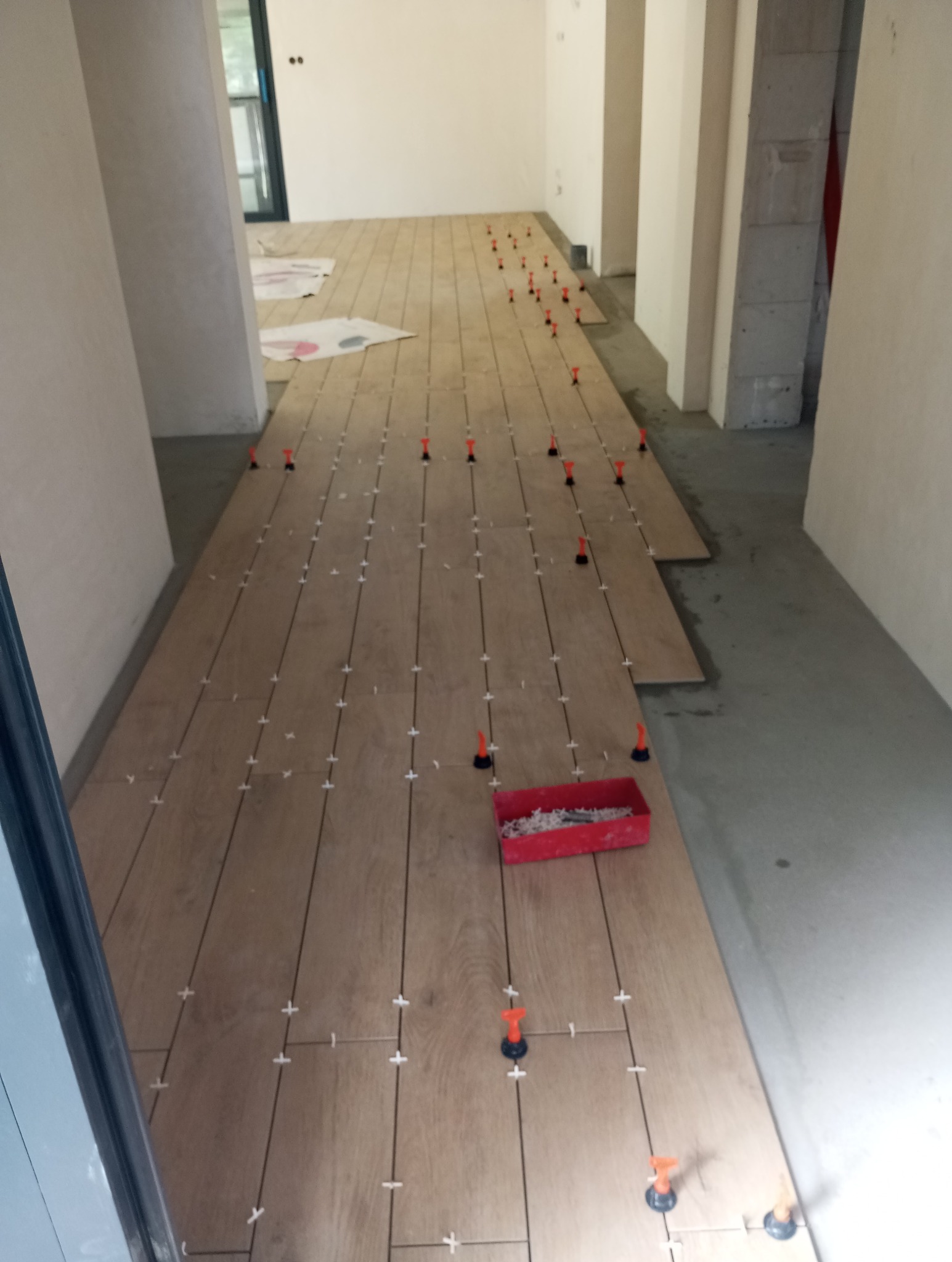 Układanie płytek imitujących drewno w korytarzu, system poziomowania płytek z czerwonymi klinami. W tle widoczne surowe ściany i przygotowane podłoże.
