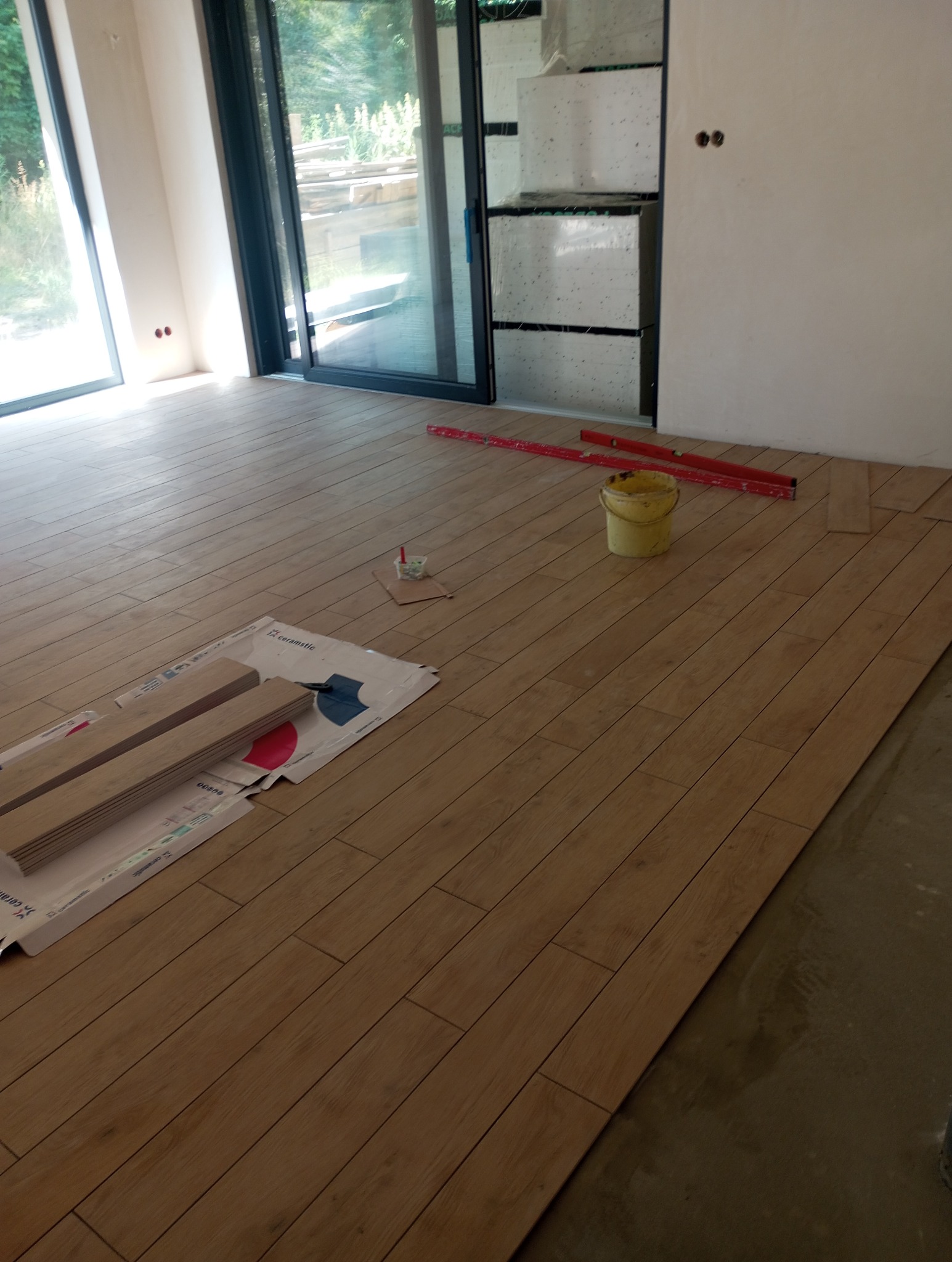 Układanie paneli podłogowych w Chorzelach. Widoczne panele, poziomica i wiadro. Częściowo ułożona podłoga z paneli drewnopodobnych, duże okno balkonowe.