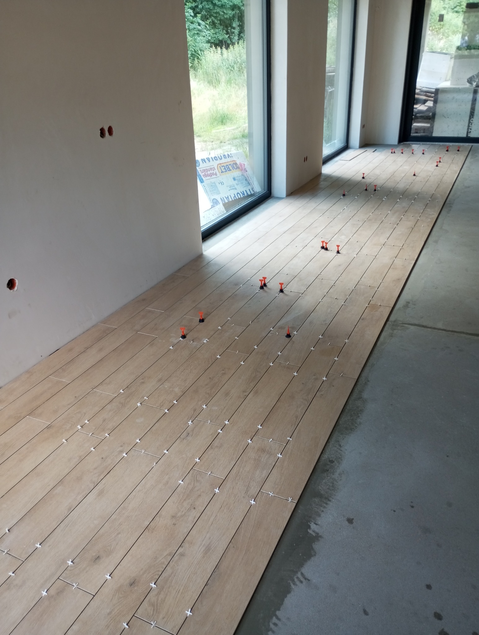 Układanie podłogi z paneli drewnopodobnych, widoczne krzyżyki dystansowe i poziomowanie. Duże okna z widokiem na zieleń. Częściowo surowy stan.