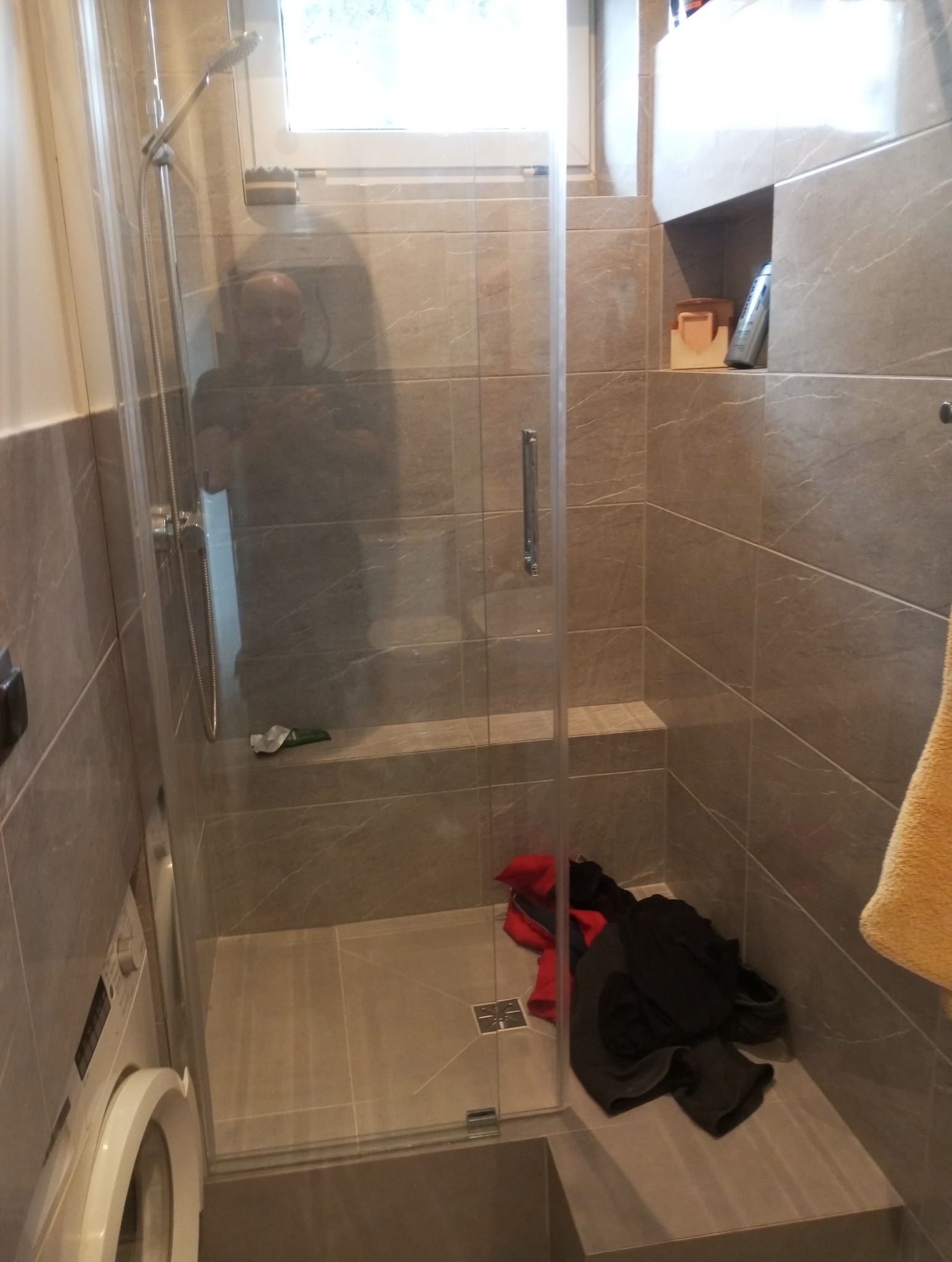 Wykończona kabina prysznicowa z szarymi płytkami imitującymi marmur, z widoczną pralką z lewej strony. W kabinie leżą ubrania.