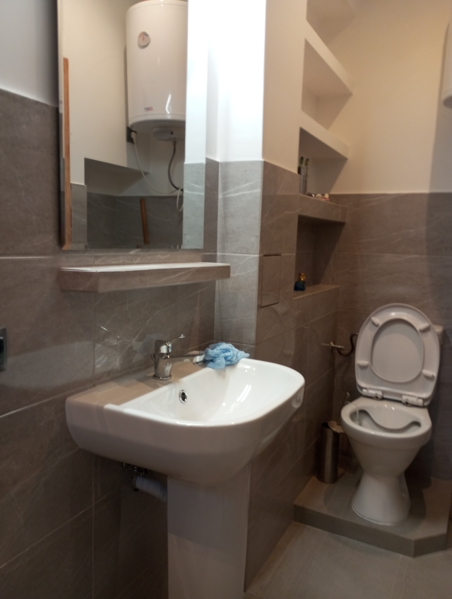 Wykończona łazienka w szarościach: umywalka na postumencie z baterią, toaleta, lustro z półką, bojler i wnęki w ścianie. Płytki imitujące marmur.