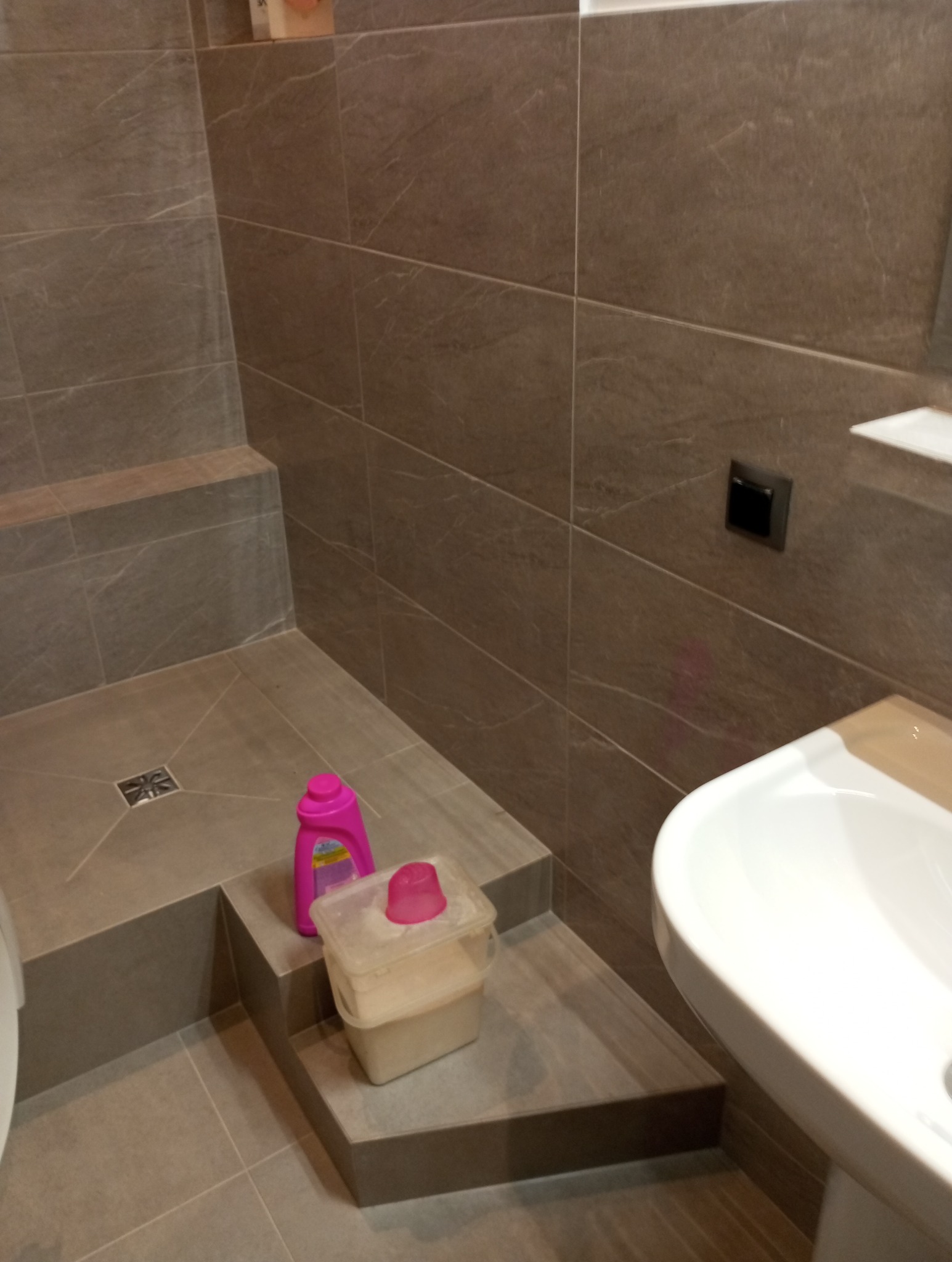 Wykończona, nowoczesna łazienka z brązowymi płytkami, umywalką i brodzikiem. Na podwyższeniu brodzika stoją środki czystości w plastikowych opakowaniach.