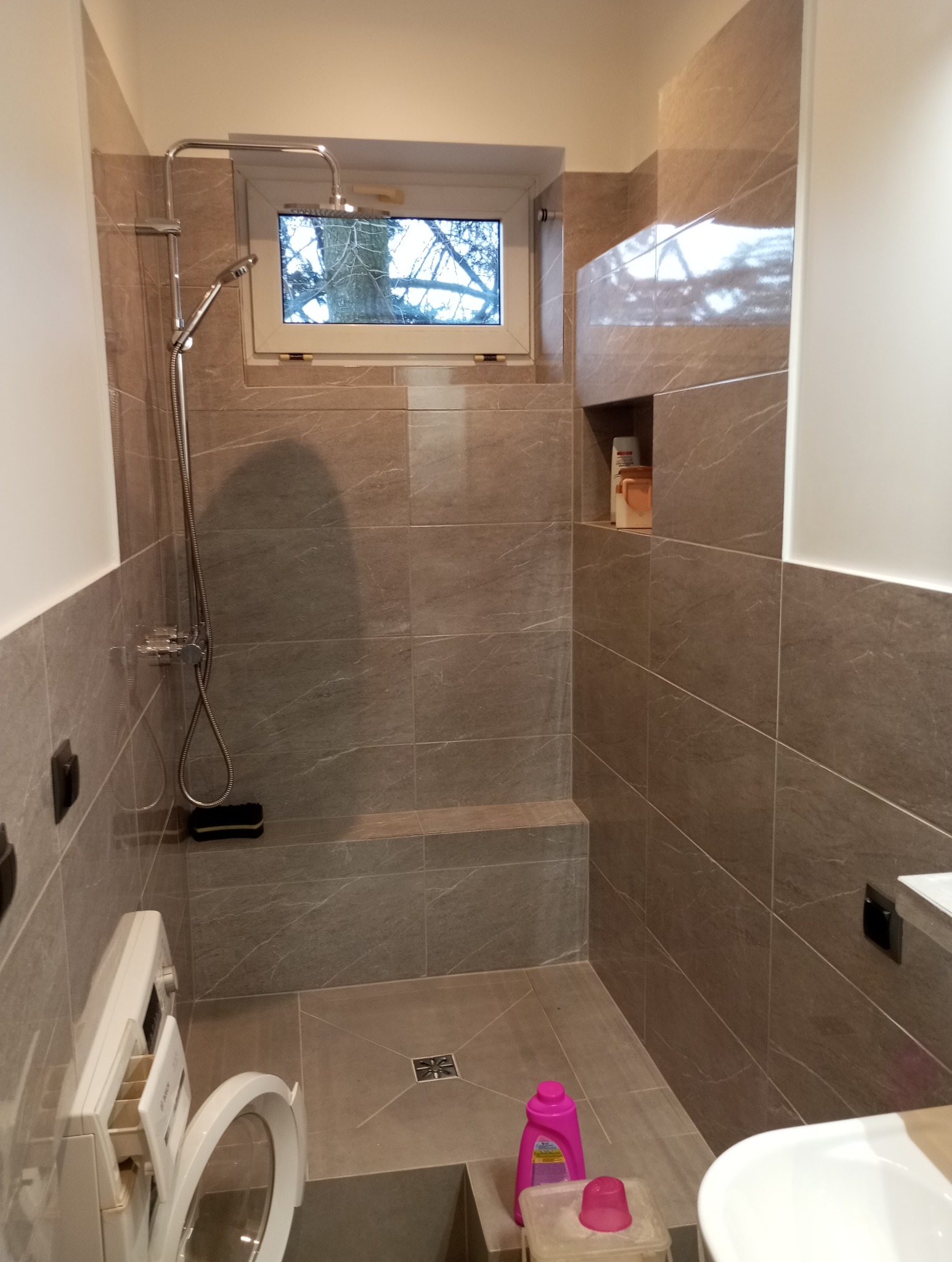 Nowoczesna kabina prysznicowa z oknem i siedziskiem, wykończona płytkami imitującymi marmur, z widoczną pralką i akcesoriami łazienkowymi.