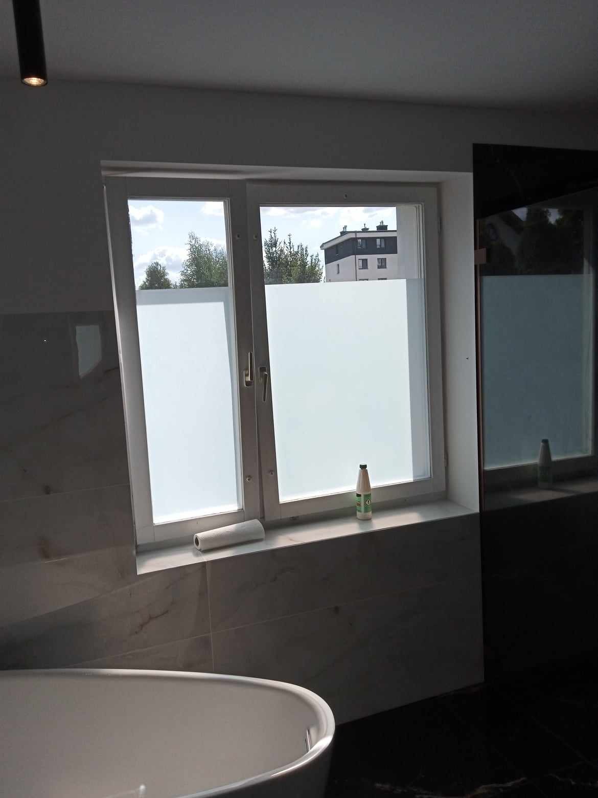 Okno w łazience z naklejoną folią matującą, częściowo widoczna wanna. Na parapecie rolka folii i butelka. Na zewnątrz widok na budynek i drzewa.