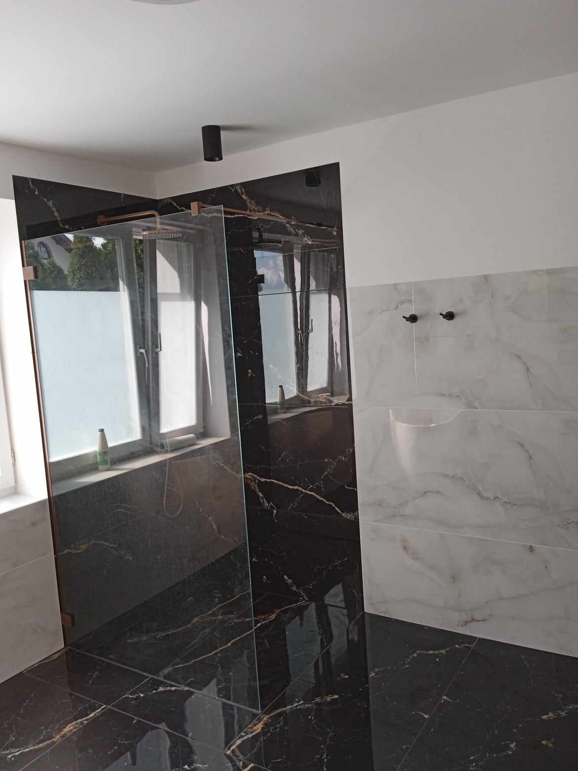 Nowoczesna łazienka z czarnymi płytkami imitującymi marmur, szklaną kabiną prysznicową i białymi płytkami na ścianie. Widoczne okno z mleczną folią.