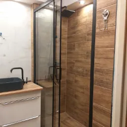 Nowoczesna łazienka z kabiną prysznicową typu walk-in z czarną ramą, drewnopodobnymi płytkami na ścianach i podłodze, oraz umywalką nablatową w kolorze grafitowym na szafce z szufladami.