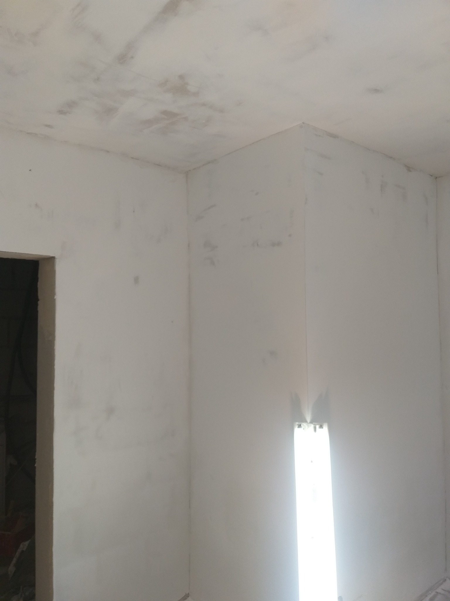 Wnętrze pokoju w trakcie prac wykończeniowych; ściany i sufit pokryte warstwą szpachli, widoczne nierówności i ślady po szpachlowaniu, w rogu pionowo zamontowana lampa halogenowa.