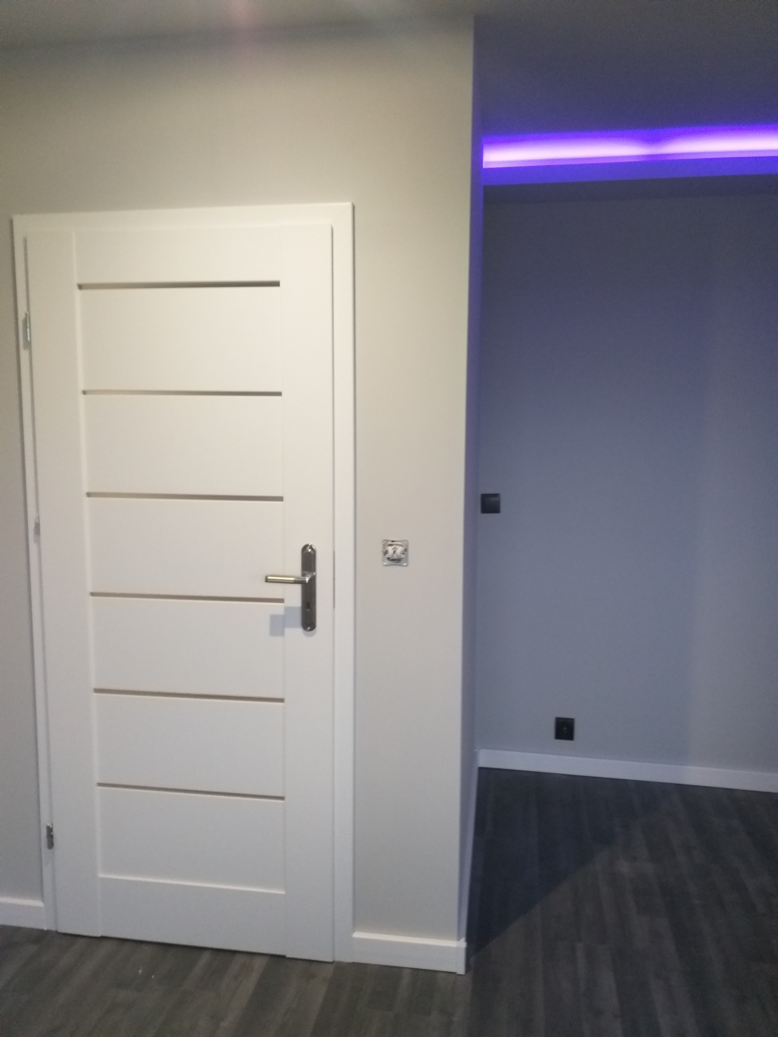 Białe, nowoczesne drzwi wewnętrzne z poziomymi frezowaniami, zamontowane w szarej ścianie, z widocznym fragmentem pomieszczenia z podświetleniem LED w kolorze fioletowym.