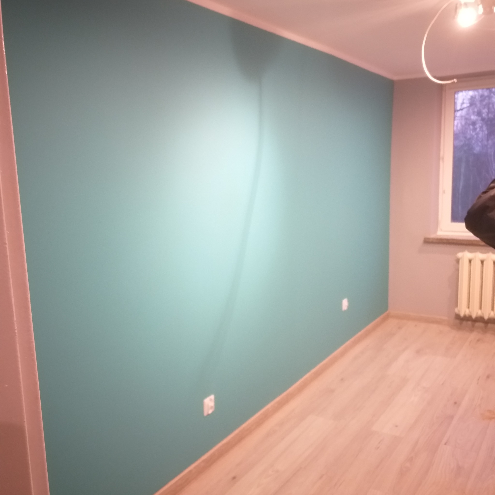 Świeżo pomalowana na turkusowo ściana w pokoju w Krasnystawie, widoczne gniazdka elektryczne, jasna podłoga z paneli, grzejnik pod oknem.