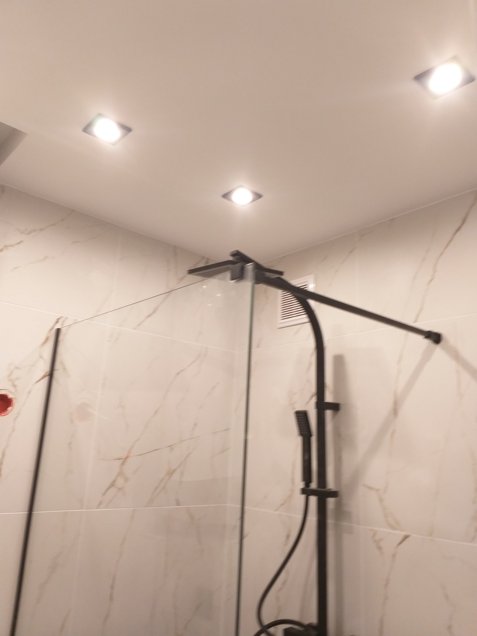 Nowoczesna kabina prysznicowa z czarną armaturą i szklaną ścianką, wykończona marmurowymi płytkami, oświetlona punktowymi lampami sufitowymi.