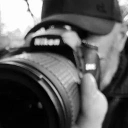 Czarno-białe ujęcie zbliżeniowe aparatu Nikon w rękach osoby w czapce z daszkiem, nieostre tło.