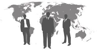 Sylwetki trzech mężczyzn w garniturach na tle szarej mapy świata. Mężczyzna pośrodku ma ręce splecione przed sobą, a dwaj po bokach mają ręce wzdłuż ciała.